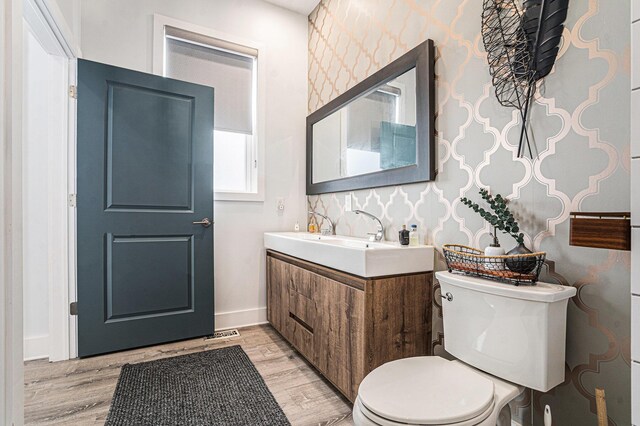 half bathroom with vanity, wood finished floors, toilet, baseboards, and wallpapered walls