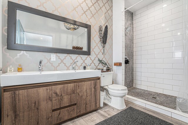 full bathroom with a tile shower, wood finished floors, vanity, toilet, and wallpapered walls
