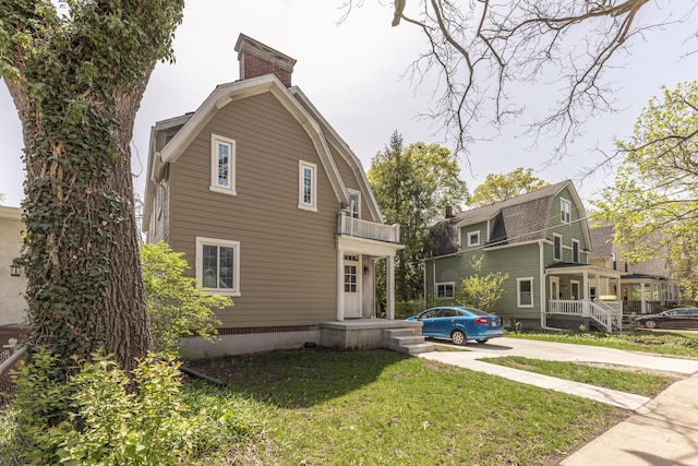 rear view of house featuring a gambrel roof, a chimney, concrete driveway, a balcony, and a lawn