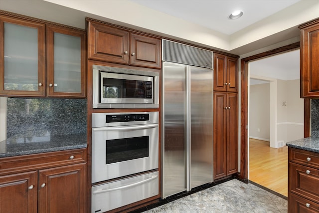 kitchen with a warming drawer, dark stone counters, tasteful backsplash, glass insert cabinets, and built in appliances