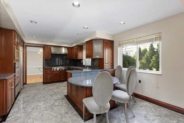 kitchen featuring a kitchen breakfast bar, wall chimney exhaust hood, stainless steel appliances, a peninsula, and glass insert cabinets