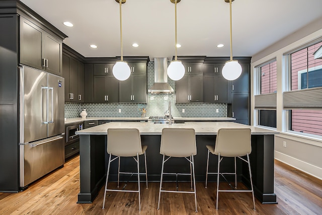 kitchen with wall chimney range hood, backsplash, a center island with sink, high quality fridge, and wood finished floors
