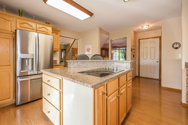 kitchen with light stone counters, black electric stovetop, stainless steel fridge with ice dispenser, light wood-style floors, and a kitchen island with sink