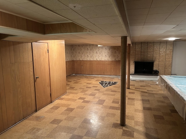 finished basement with wood walls, wainscoting, a brick fireplace, light floors, and a drop ceiling