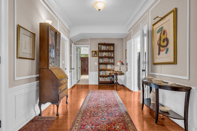 corridor featuring a decorative wall, wood finished floors, crown molding, and a wainscoted wall