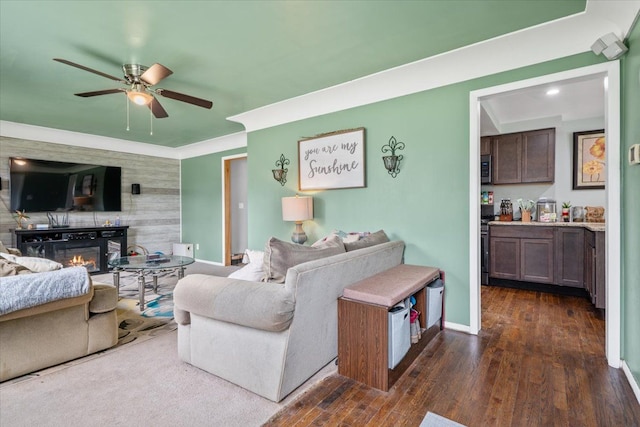 living area featuring a glass covered fireplace, baseboards, a ceiling fan, and dark wood finished floors