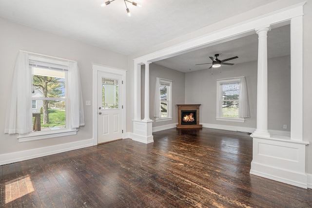 unfurnished living room with a warm lit fireplace, wood-type flooring, a ceiling fan, baseboards, and decorative columns