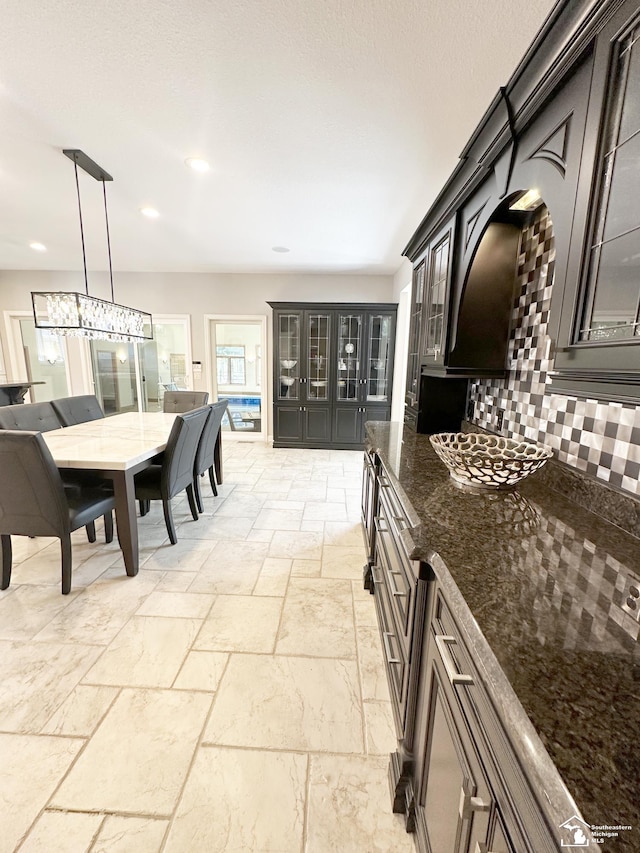 kitchen featuring stone tile floors, dark stone counters, decorative backsplash, pendant lighting, and glass insert cabinets