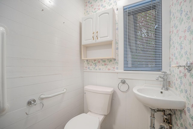 half bathroom with a sink, wainscoting, toilet, and wallpapered walls