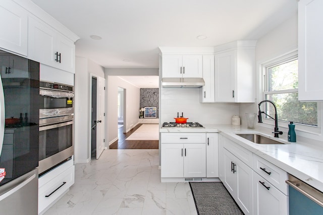 kitchen with under cabinet range hood, white cabinets, light stone counters, a sink, and appliances with stainless steel finishes