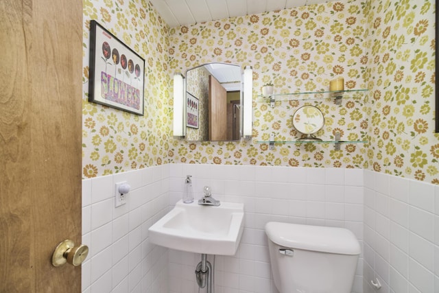 bathroom featuring tile walls, wainscoting, a sink, toilet, and wallpapered walls