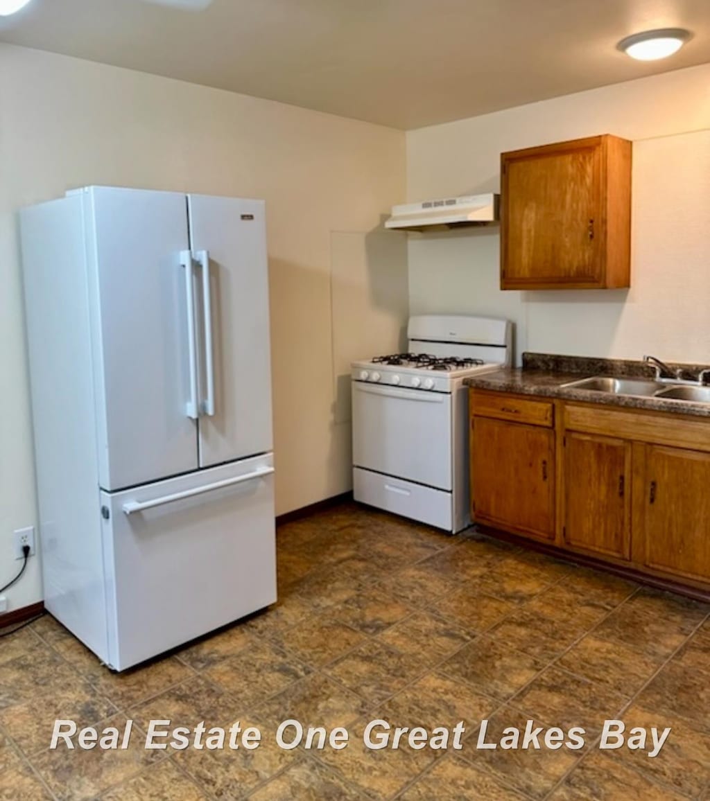 kitchen featuring dark countertops, brown cabinetry, under cabinet range hood, and white appliances
