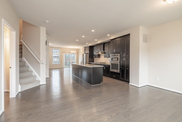 kitchen with an inviting chandelier, a kitchen island with sink, recessed lighting, baseboards, and dark wood-type flooring