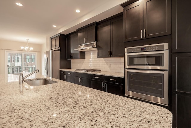 kitchen featuring under cabinet range hood, light stone countertops, a sink, a chandelier, and stainless steel appliances