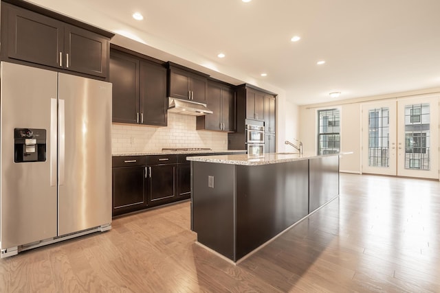 kitchen with appliances with stainless steel finishes, light wood finished floors, a kitchen island with sink, decorative backsplash, and under cabinet range hood