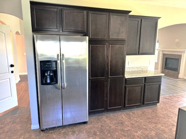 kitchen featuring arched walkways, a fireplace, dark brown cabinets, stainless steel fridge with ice dispenser, and light stone counters