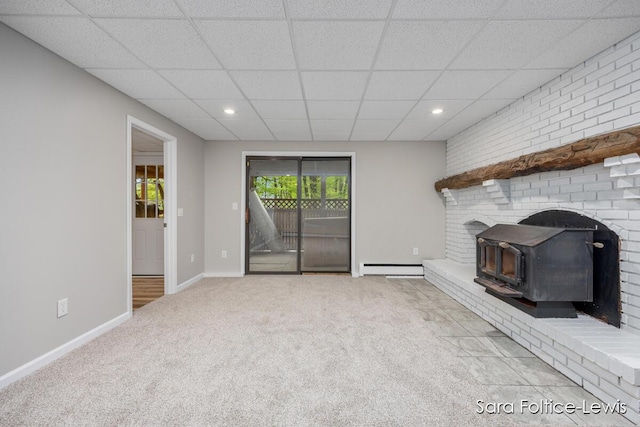 unfurnished living room with carpet flooring, a drop ceiling, baseboards, a baseboard heating unit, and a wood stove
