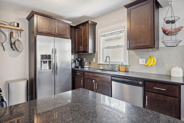kitchen with dark brown cabinets, appliances with stainless steel finishes, a sink, dark stone counters, and decorative backsplash