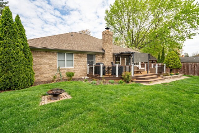 back of house featuring a fire pit, a wooden deck, roof with shingles, brick siding, and a lawn