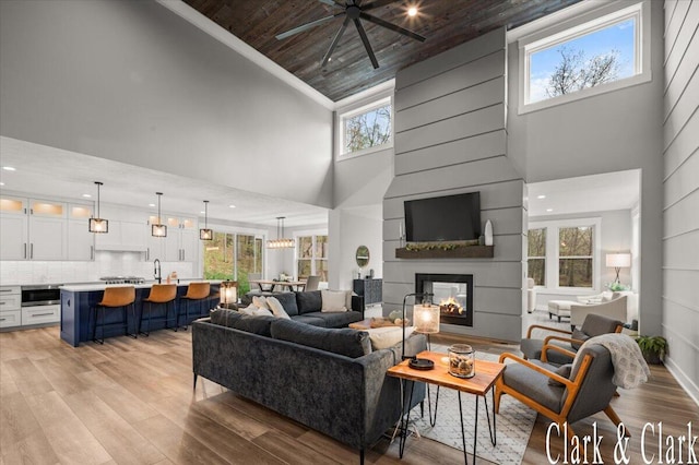 living room featuring ceiling fan, high vaulted ceiling, wooden ceiling, a large fireplace, and light wood finished floors