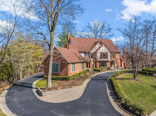 tudor-style house with a chimney, roof with shingles, a front yard, brick siding, and driveway