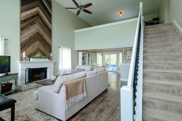 living room featuring stairway, high vaulted ceiling, ceiling fan, a fireplace with raised hearth, and wood finished floors