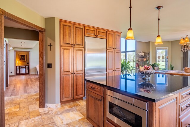 kitchen with built in appliances, brown cabinets, dark countertops, decorative light fixtures, and light stone finish flooring