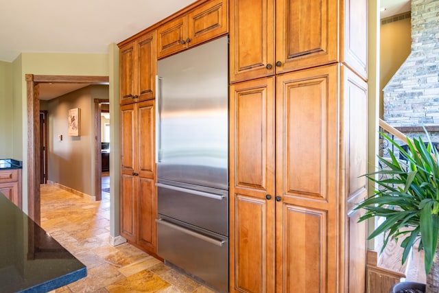 kitchen featuring built in refrigerator, light stone finish floors, brown cabinets, and dark stone countertops