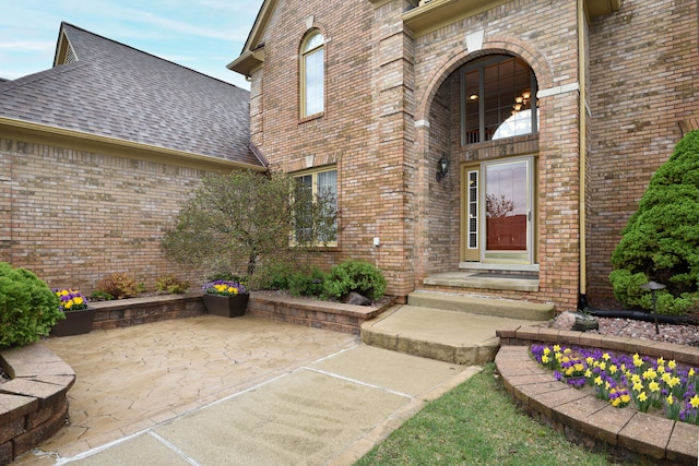 doorway to property featuring brick siding, roof with shingles, a patio, and uncovered parking