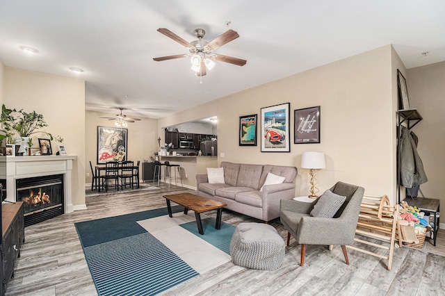 living area with light wood-style flooring, a ceiling fan, and a glass covered fireplace