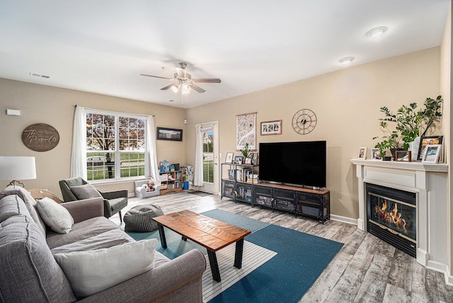 living room with wood finished floors, a ceiling fan, visible vents, baseboards, and a glass covered fireplace