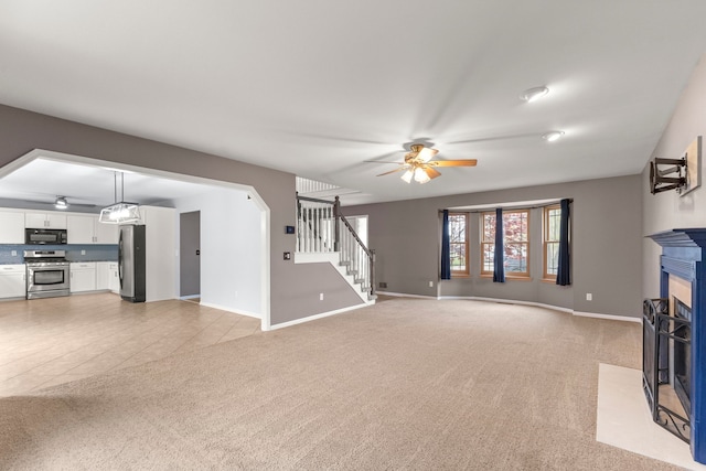 unfurnished living room with a ceiling fan, a fireplace with flush hearth, baseboards, light carpet, and stairs