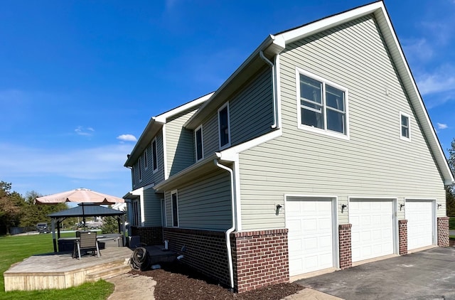 view of side of home with an attached garage, a wooden deck, a gazebo, brick siding, and driveway