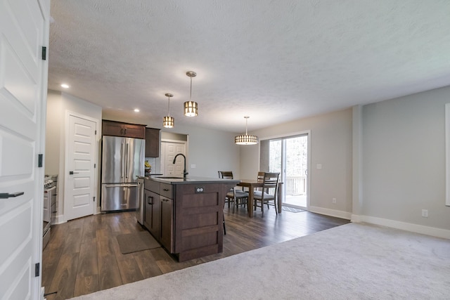 kitchen featuring an island with sink, a sink, freestanding refrigerator, dark brown cabinetry, and baseboards
