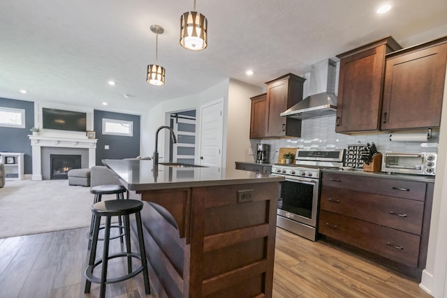 kitchen with a sink, dark countertops, a kitchen island with sink, gas range, and wall chimney range hood