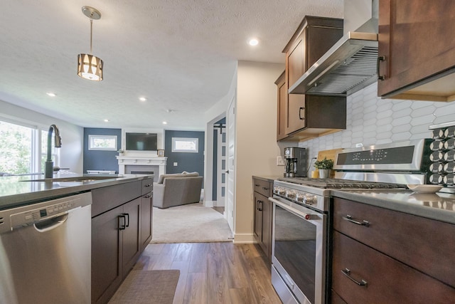 kitchen featuring a sink, stainless steel appliances, wall chimney exhaust hood, a fireplace, and dark wood-style floors
