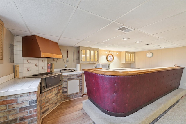 interior space with a drop ceiling, tile countertops, visible vents, wall chimney range hood, and light wood-style floors