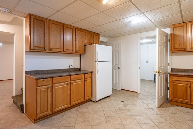 kitchen featuring freestanding refrigerator, dark countertops, brown cabinets, a drop ceiling, and light tile patterned flooring