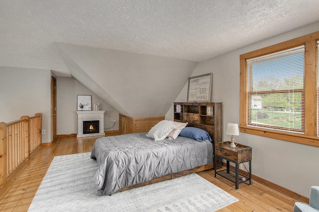 bedroom featuring lofted ceiling, a warm lit fireplace, a textured ceiling, baseboards, and hardwood / wood-style floors