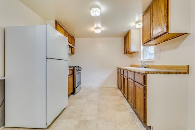 kitchen featuring freestanding refrigerator, stainless steel gas range oven, a sink, brown cabinetry, and light countertops