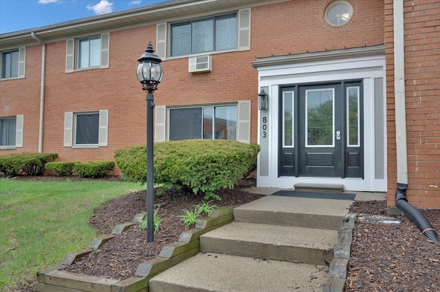 view of exterior entry with brick siding, a wall unit AC, and a lawn
