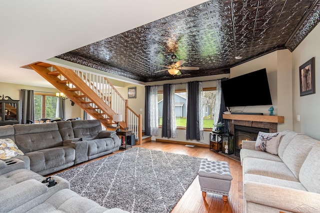 living area with stairs, a warm lit fireplace, an ornate ceiling, wood finished floors, and ceiling fan