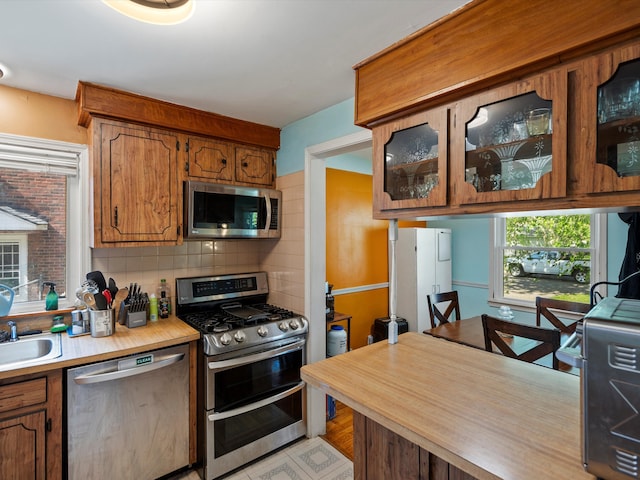 kitchen featuring appliances with stainless steel finishes, light countertops, brown cabinets, decorative backsplash, and glass insert cabinets