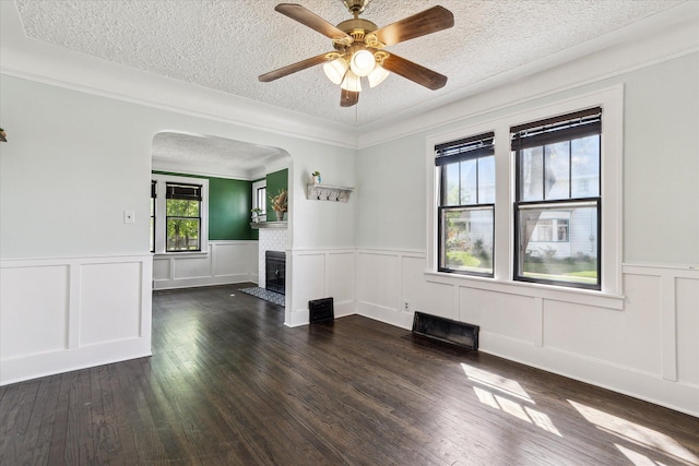 empty room featuring a textured ceiling, wainscoting, dark wood-type flooring, arched walkways, and a fireplace