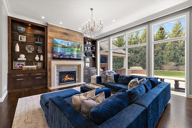 living room with a warm lit fireplace, dark wood-style floors, a chandelier, built in shelves, and recessed lighting