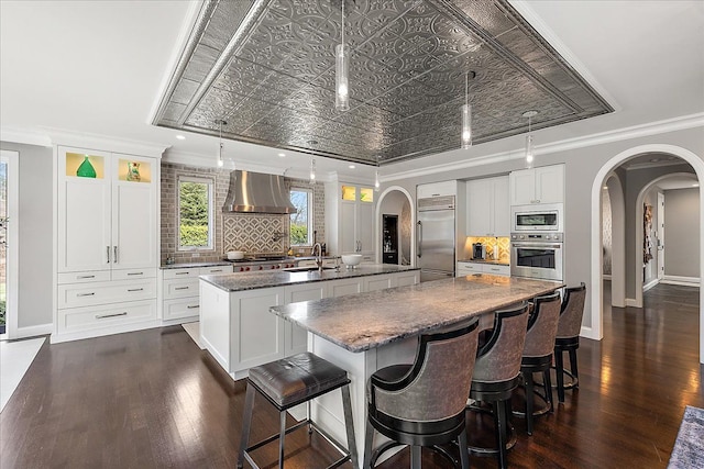 kitchen featuring a large island with sink, built in appliances, arched walkways, wall chimney exhaust hood, and an ornate ceiling