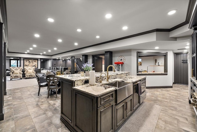kitchen with ornamental molding, a sink, a kitchen island with sink, baseboards, and light stone counters