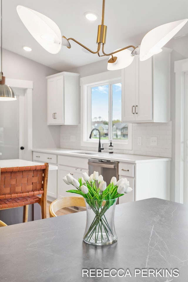 kitchen featuring backsplash, white cabinets, stainless steel dishwasher, a sink, and decorative light fixtures