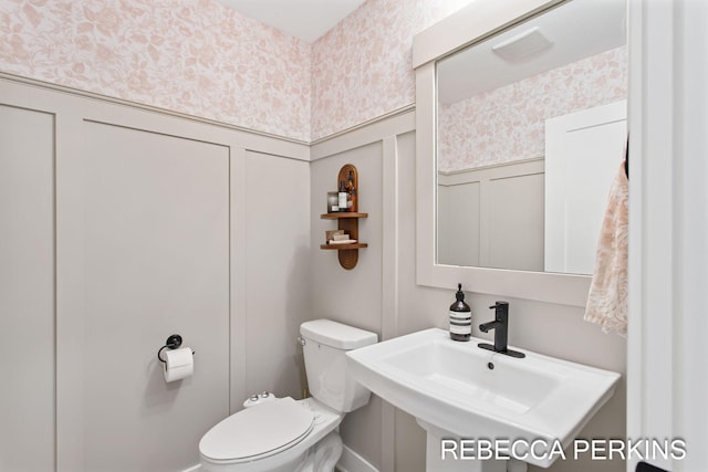bathroom with a decorative wall, wallpapered walls, toilet, a wainscoted wall, and a sink