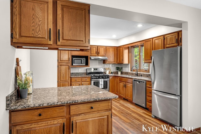 kitchen with stainless steel appliances, under cabinet range hood, light wood-style floors, a peninsula, and brown cabinetry